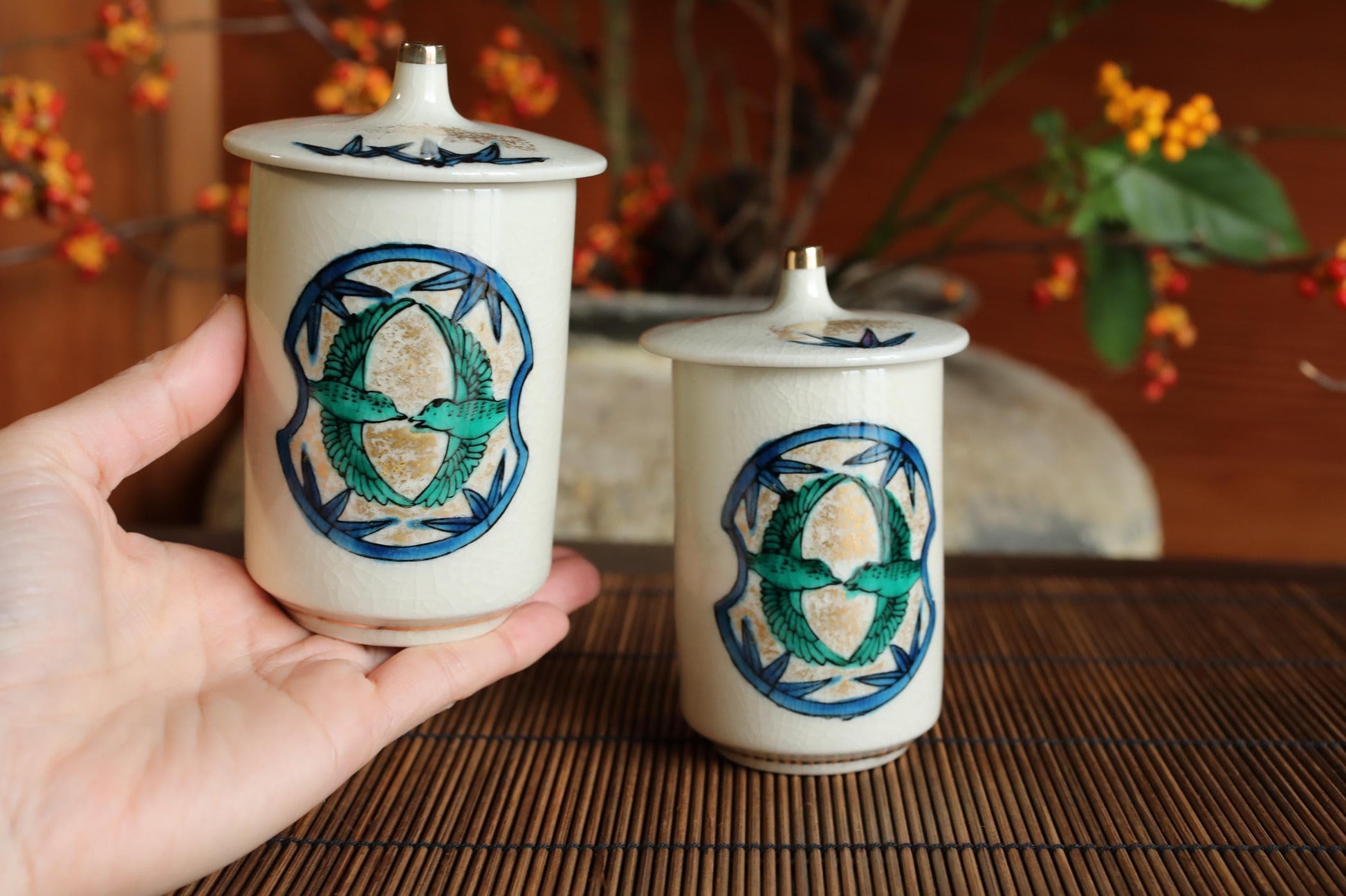 A hand is holding two white ceramic containers with blue and green designs on them, placed on a bamboo mat.