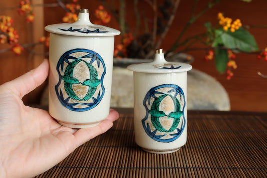 A hand is holding two white ceramic containers with blue and green designs on them, placed on a bamboo mat.
