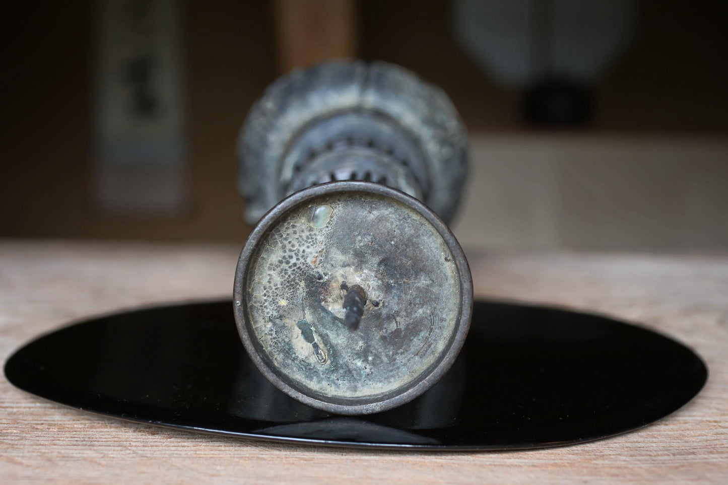 An old, rusted metal vase with a circular base sits on a black circular plate on a wooden surface.
