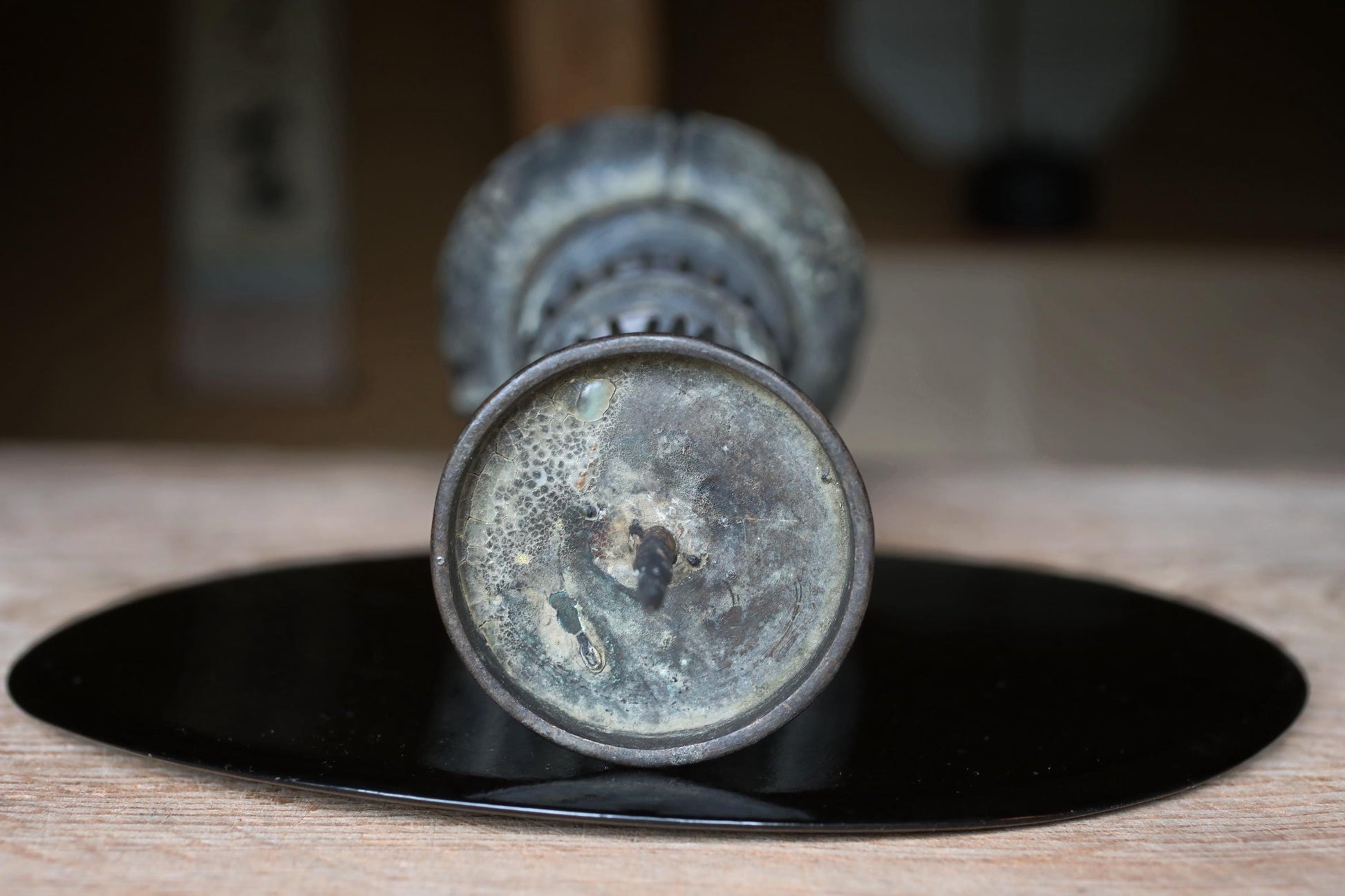 An old, rusted metal vase with a circular base sits on a black circular plate on a wooden surface.