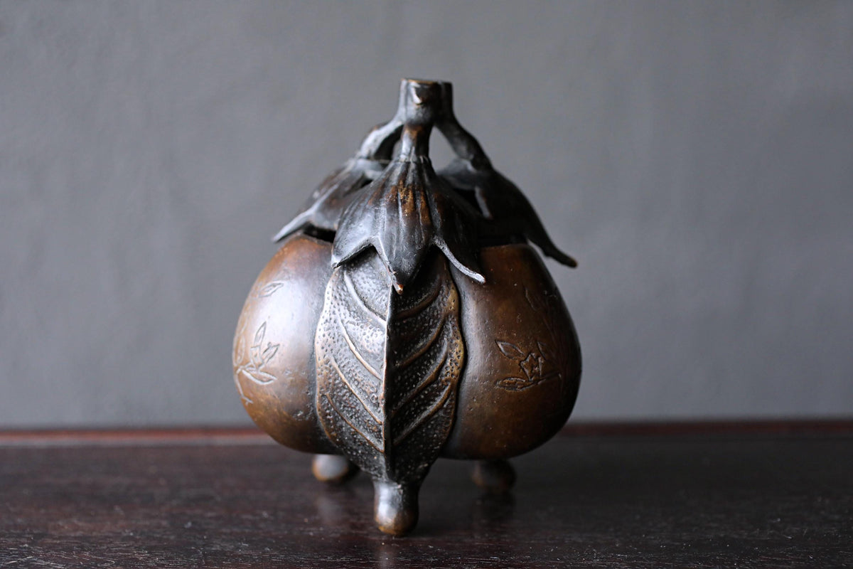 A bronze incense burner with a leaf-like design on the front sits on a dark surface.