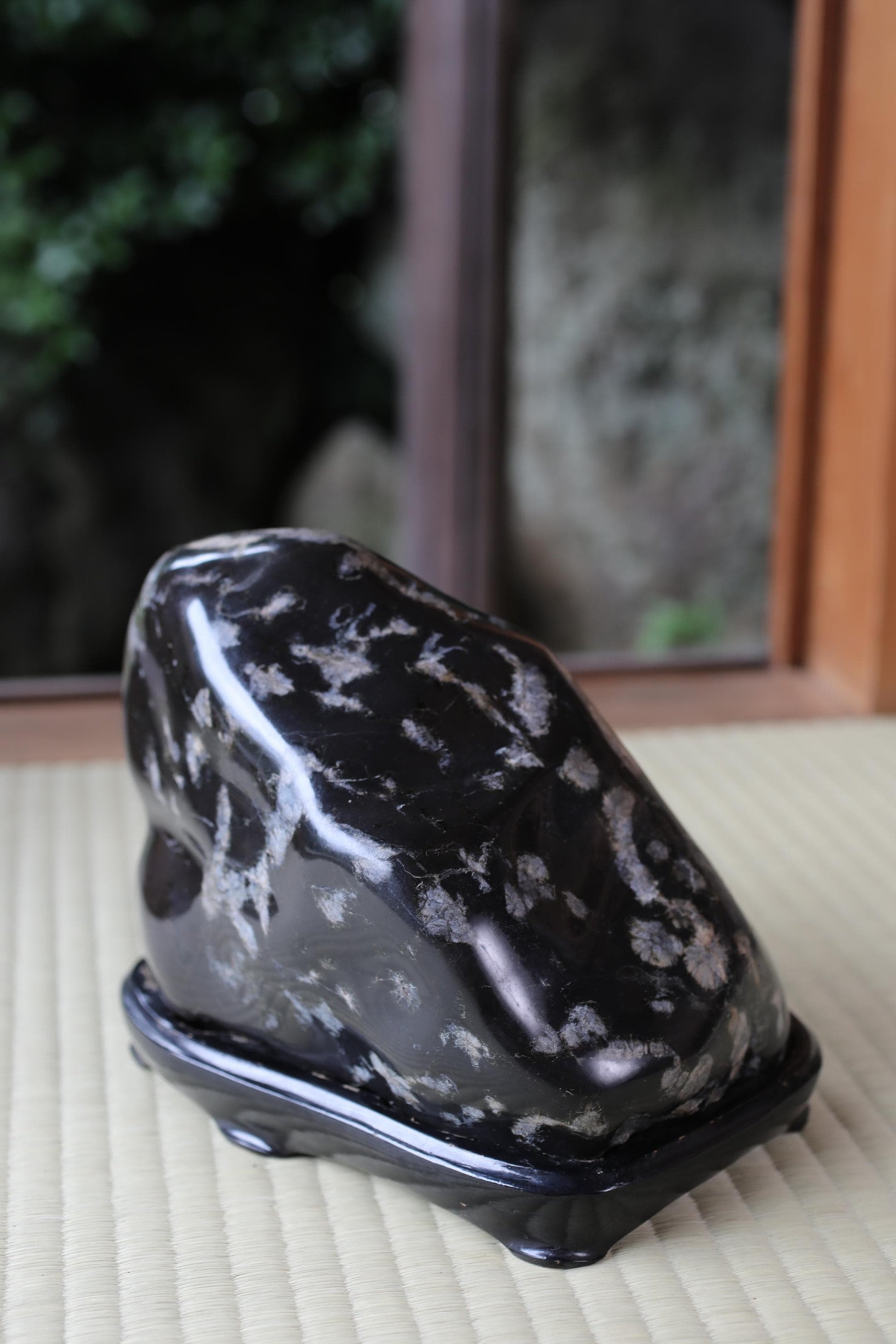 A black, speckled rock sits on a black stand, positioned in front of a window with a stone wall visible.