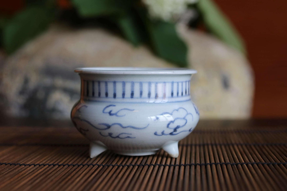 A white ceramic bowl with blue and white designs sits on a bamboo mat, with a blurred background of green leaves.