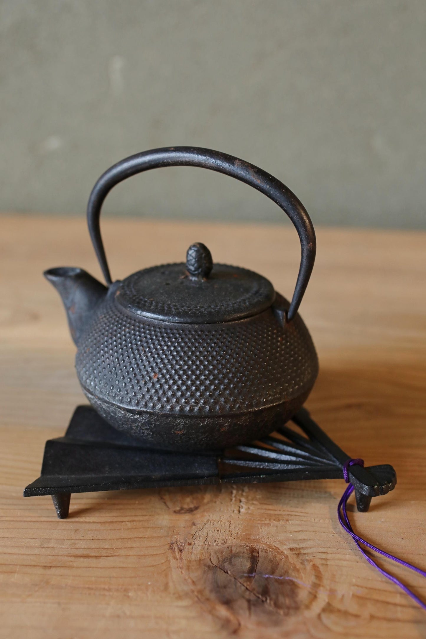 A black metal teapot with a curved handle and spout sits on a wooden surface, accompanied by a purple string.