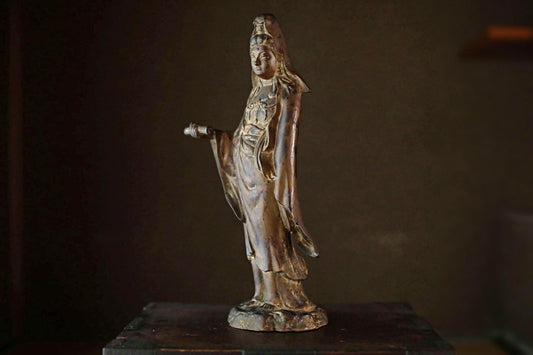 A bronze statue of a woman with a long, flowing robe and a headdress, standing on a wooden surface.