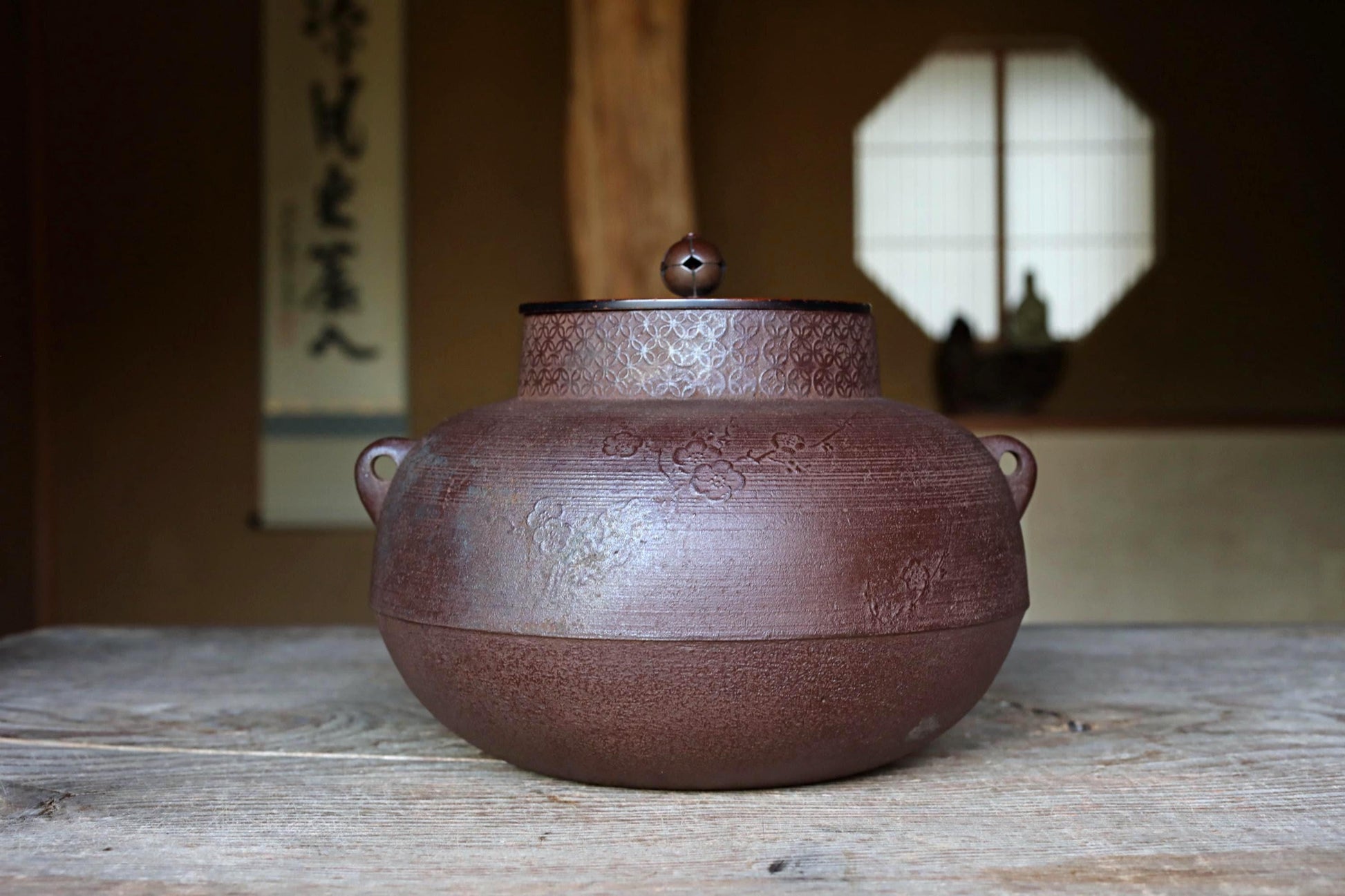 A large, round, brown ceramic teapot with a lid sits on a wooden surface, with a window and a sign with Japanese characters in the background.