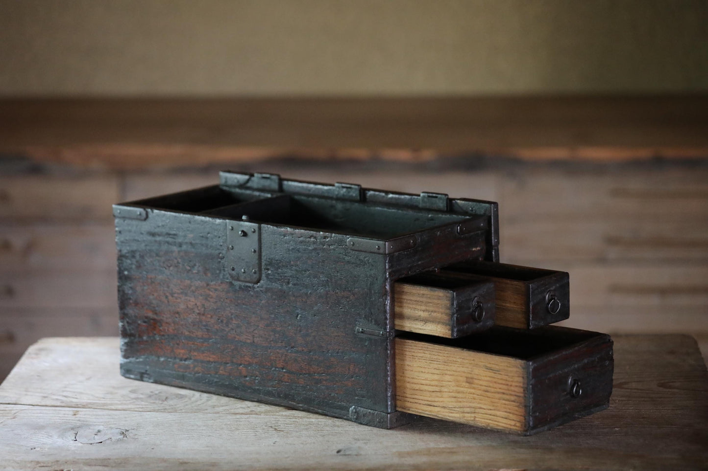 An old, weathered wooden chest with three drawers sits on a wooden surface, its metal clasps and handles showing signs of age and use.