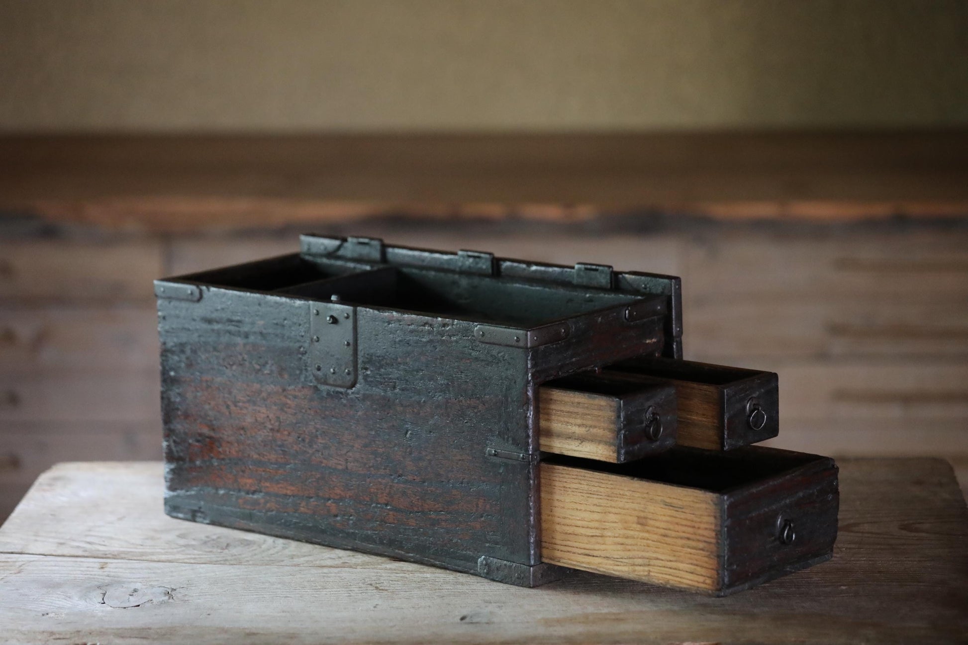An old, weathered wooden chest with three drawers sits on a wooden surface, its metal clasps and handles showing signs of age and use.