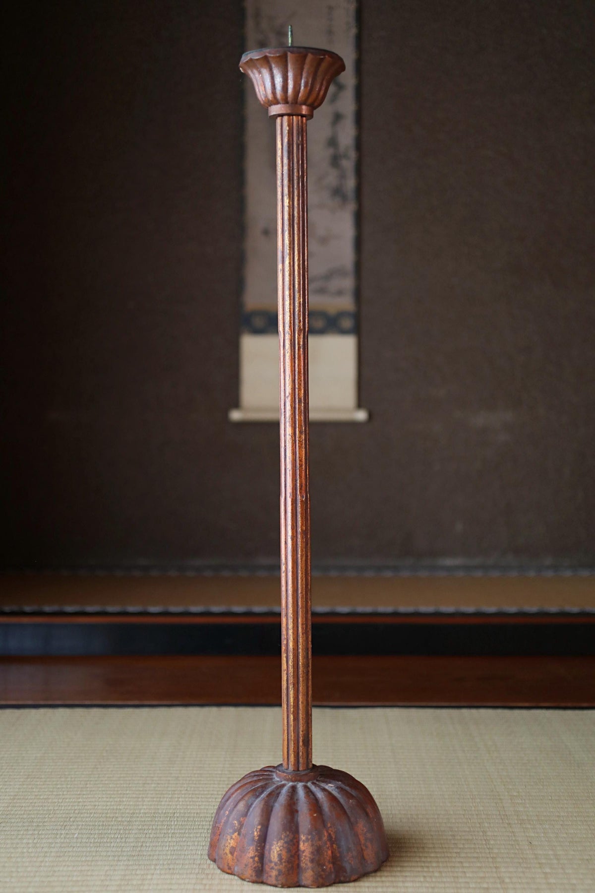 A tall, ornate candle holder with a fluted design stands on a beige mat in a dimly lit room.