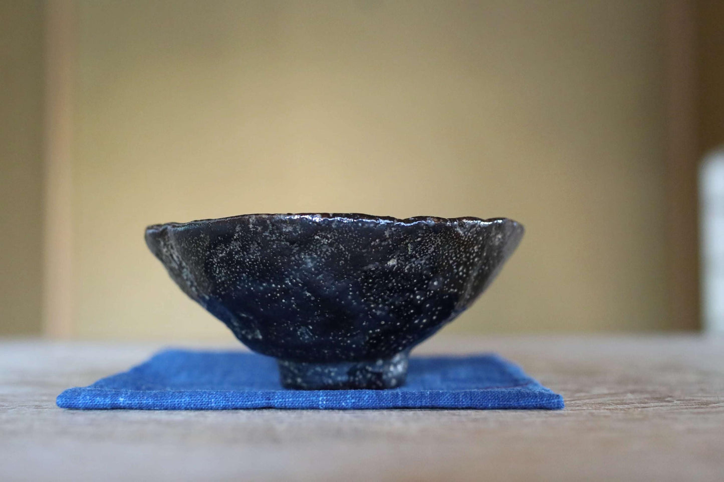 A black ceramic bowl with a speckled texture sits on a blue cloth on a wooden surface.
