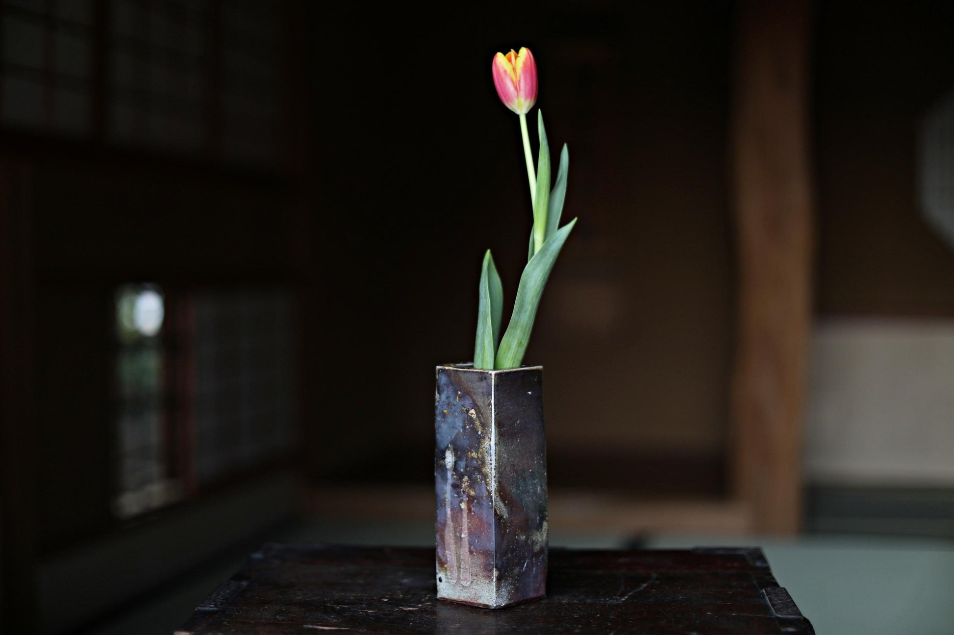 以下が含まれることがあります： 濃い灰色のセラミックの花瓶に、黄色と赤のチューリップが1本挿してあります。花瓶は表面がざらざらしていて、暗い木製のテーブルの上に置かれています。