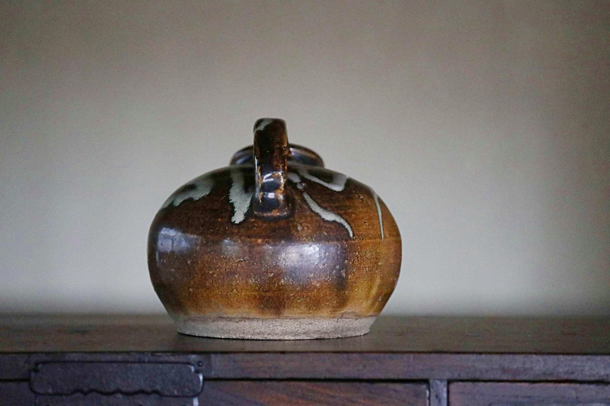 A brown ceramic jug with a handle and spout sits on a wooden surface against a plain background.