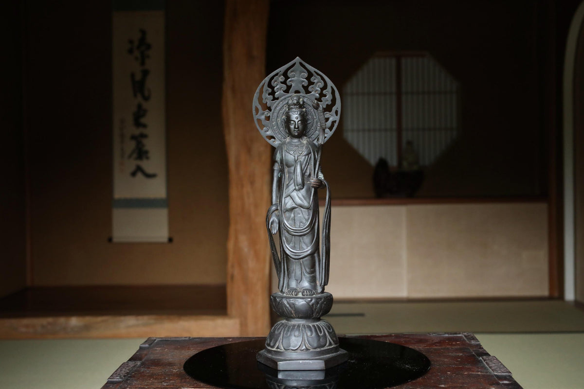 A silver statue of a deity, possibly a Buddhist or Hindu deity, stands on a wooden base in a dimly lit room with a window and a banner with Japanese characters in the background.