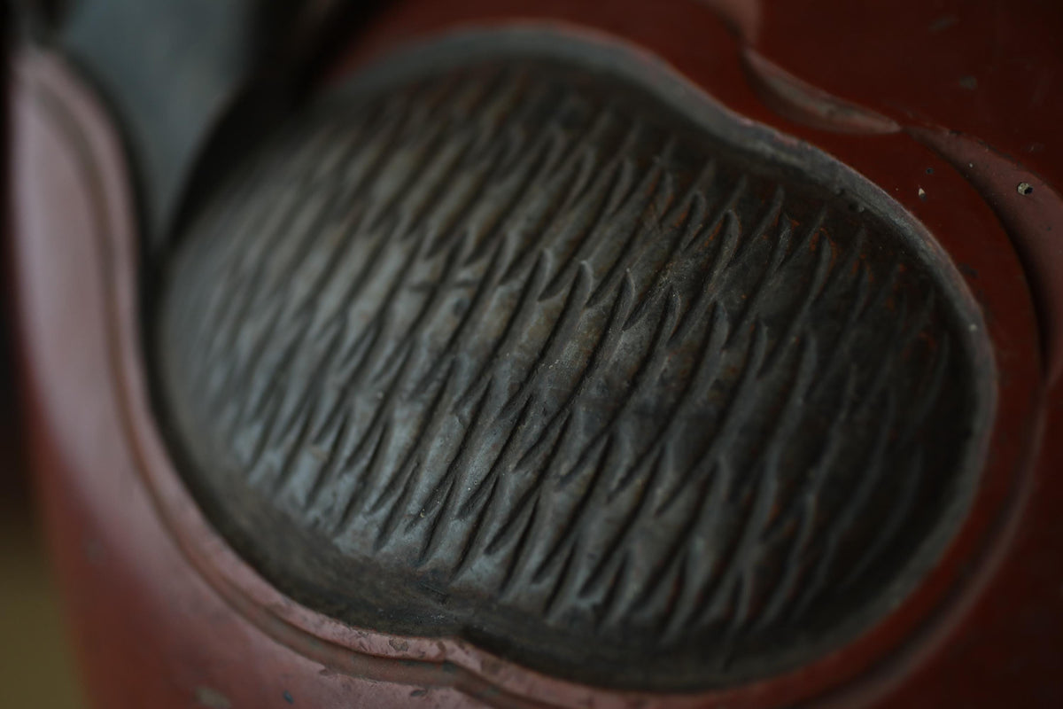 A close-up view of a textured, dark-colored surface with a pattern of lines and grooves, resembling a metal or stone texture.