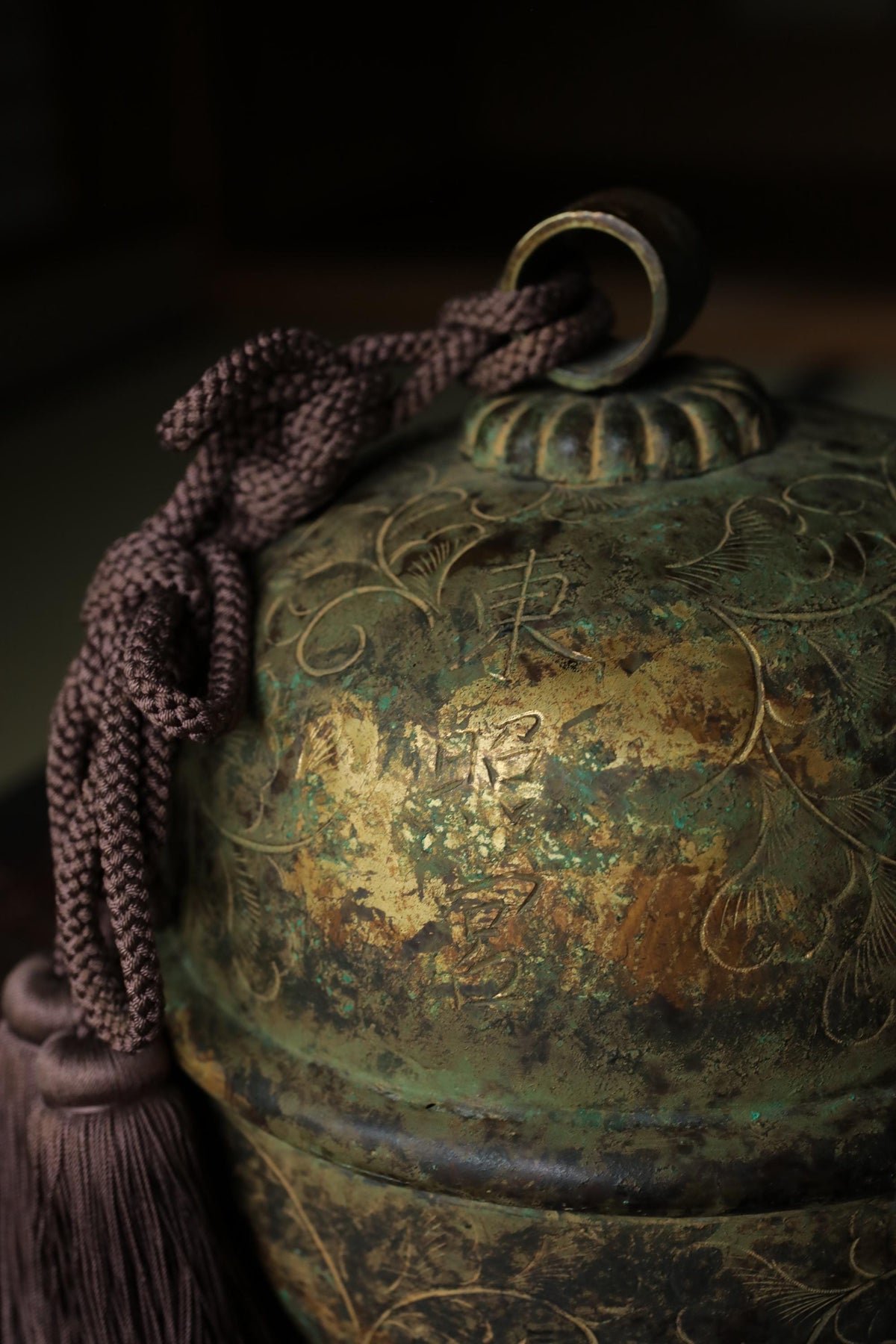 An ornate, antique-looking metal bell or container with a tassel hanging from it, set against a dark background.