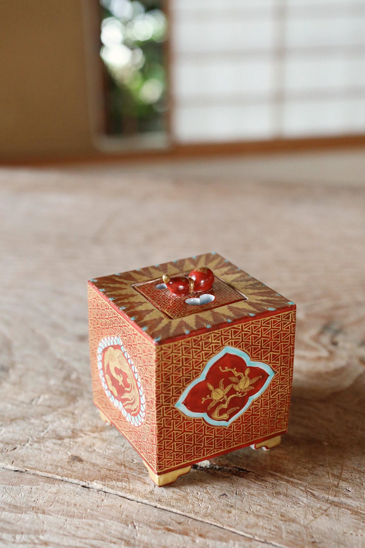 A small, ornate box with a red and gold design sits on a wooden surface, featuring a dragon motif and a red gemstone on top.