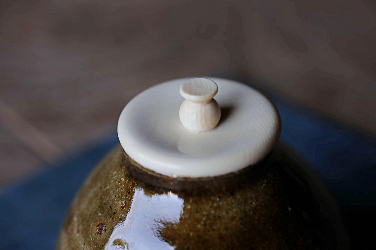 A close-up image of a ceramic jar or container with a white lid, featuring a small white knob or handle on top.