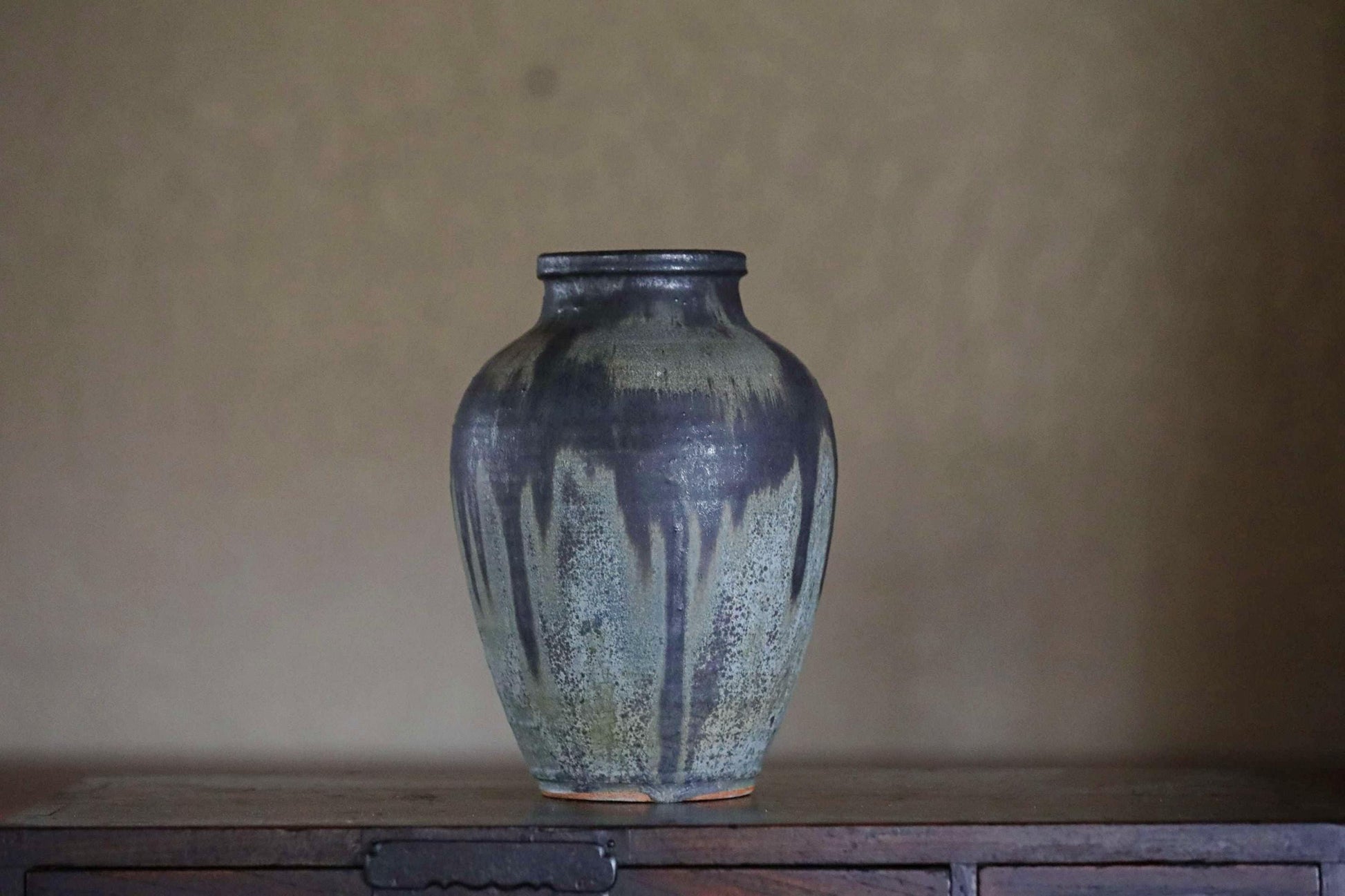 A dark blue ceramic vase with a textured surface sits on a wooden surface against a plain background.
