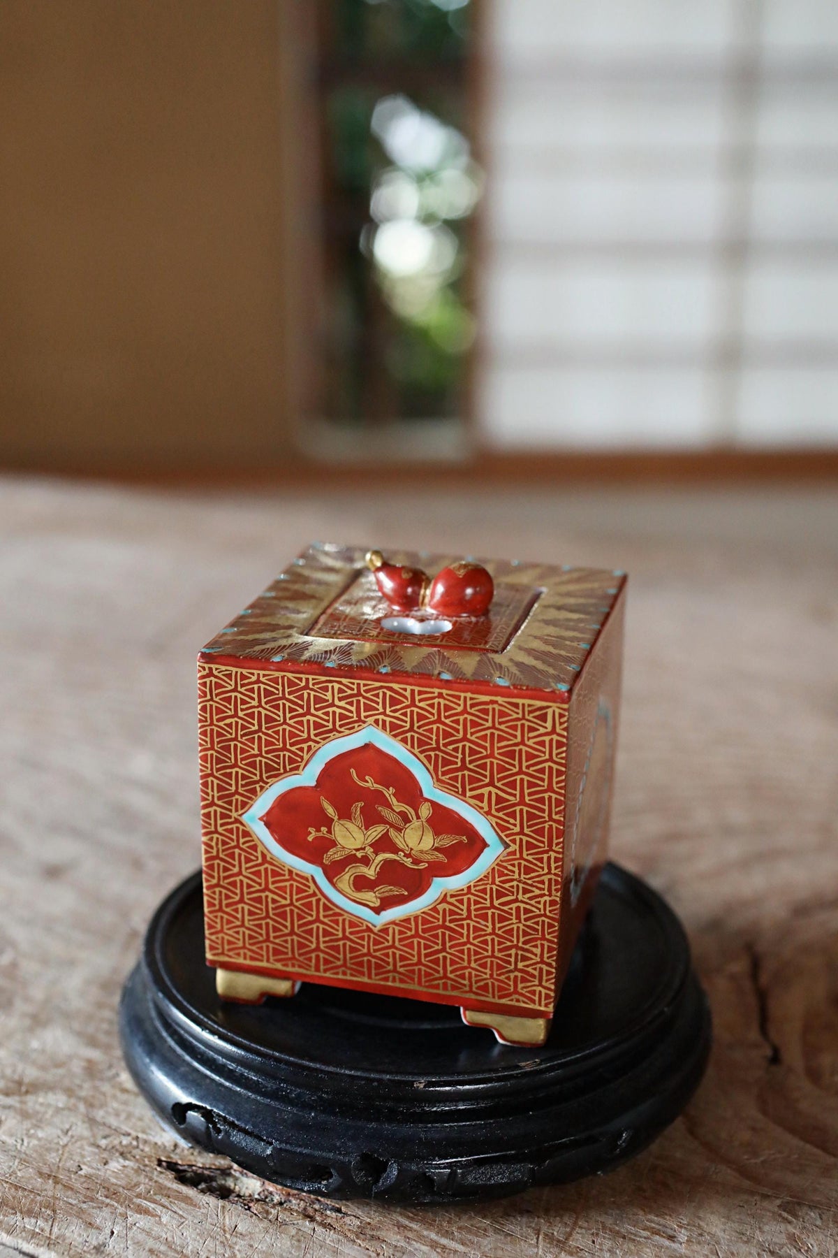 A small, ornate red and gold box with a dragon design sits on a black stand, placed on a wooden surface.