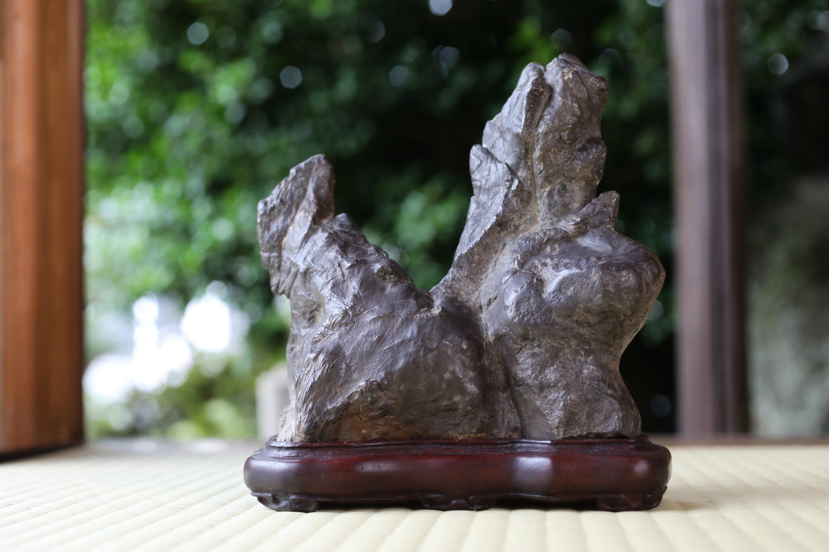 A stone sculpture of a rock formation is displayed on a wooden base, placed on a table with a blurred natural background.