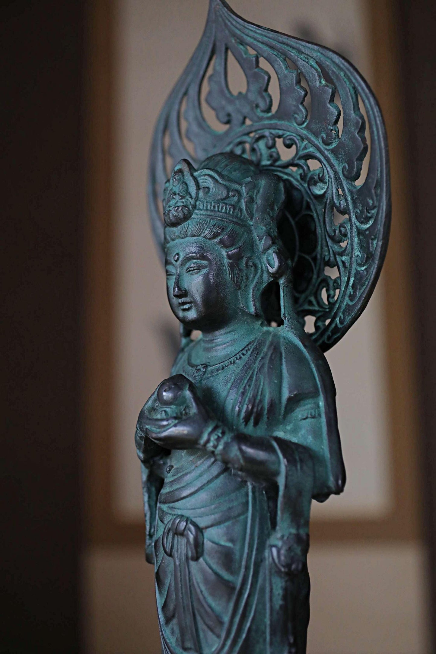A bronze statue of a deity, possibly a Buddha, with intricate designs and a flowing robe, standing in a room with a blurred background.