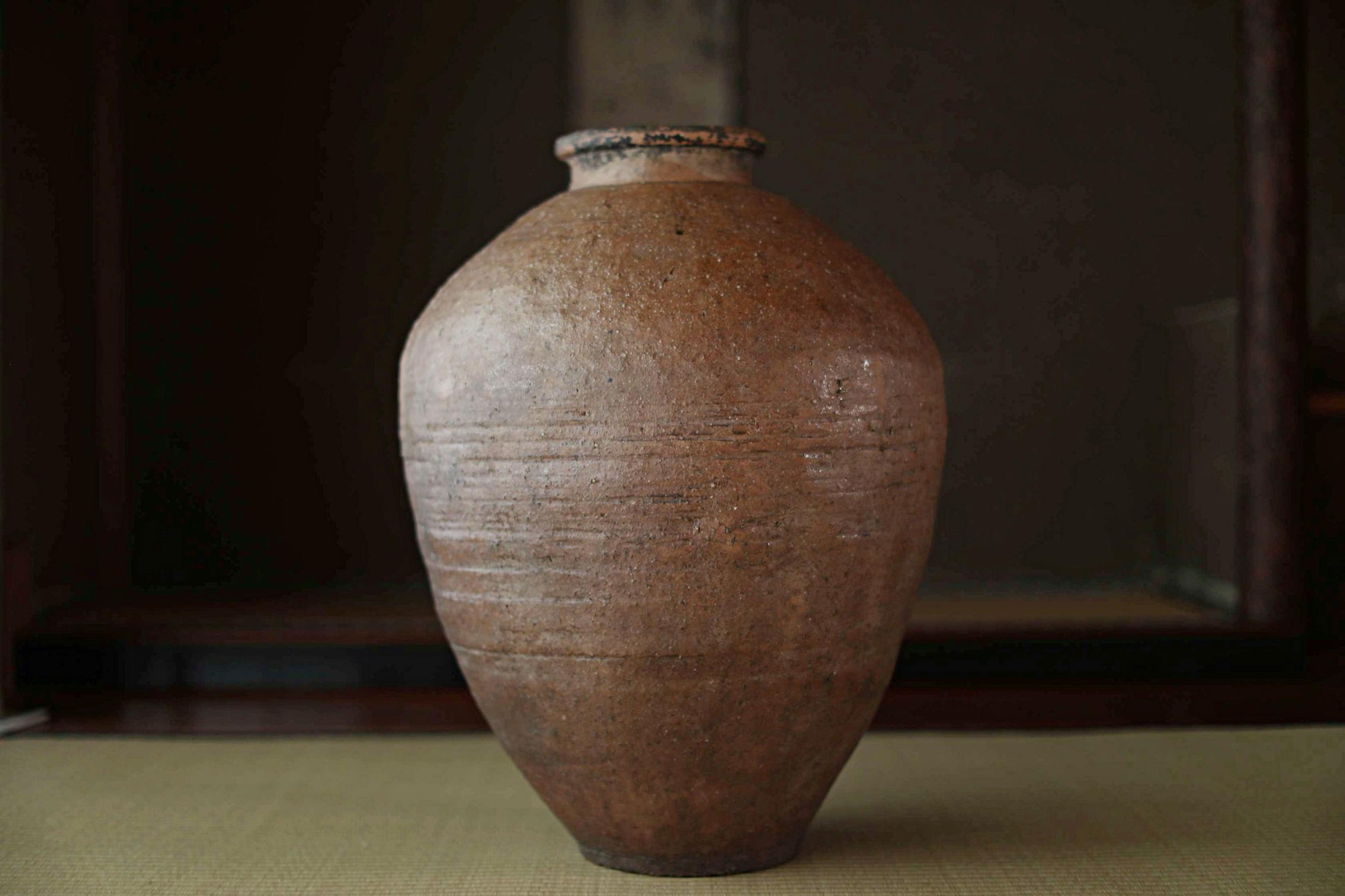 A large, brown ceramic vase with a textured surface sits on a wooden surface against a dark background.