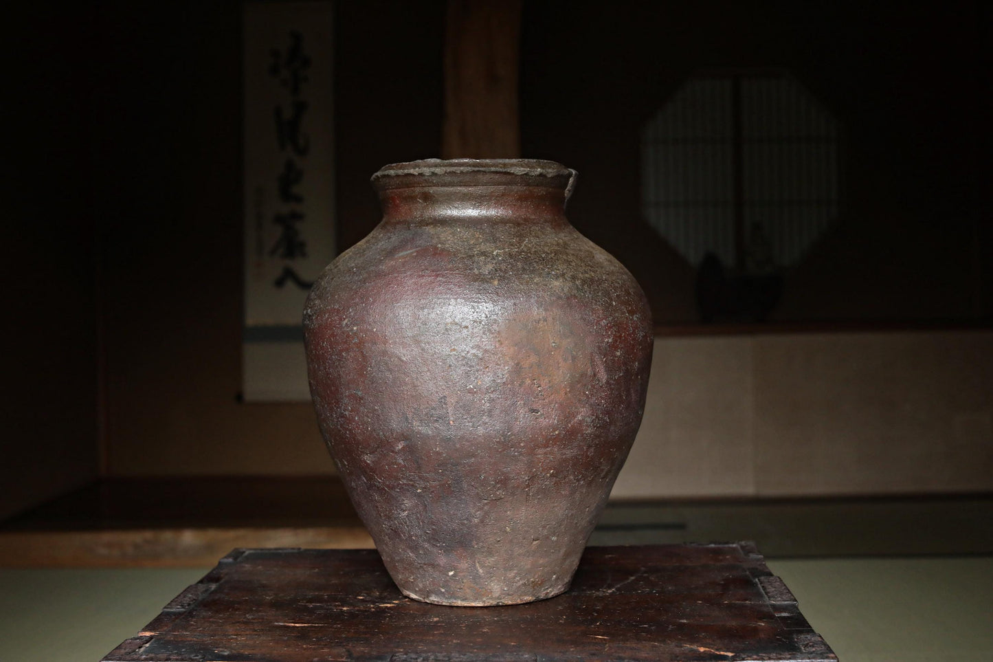 An old, weathered clay vase sits on a wooden surface, with a window and a wall with Japanese characters in the background.