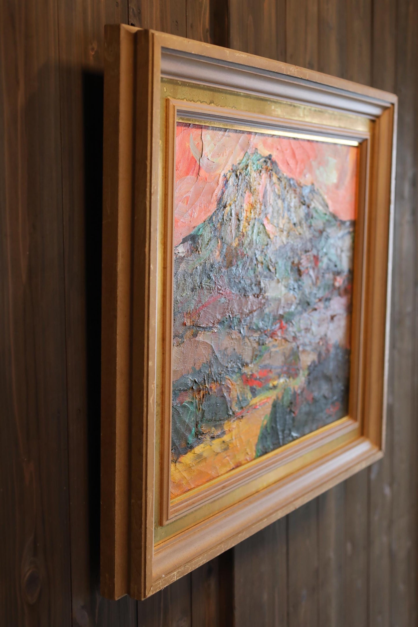 a framed painting of a mountain landscape, hanging on a wooden wall.