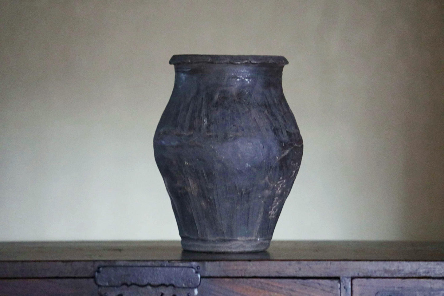 A dark-colored vase sits on a wooden surface against a plain background.