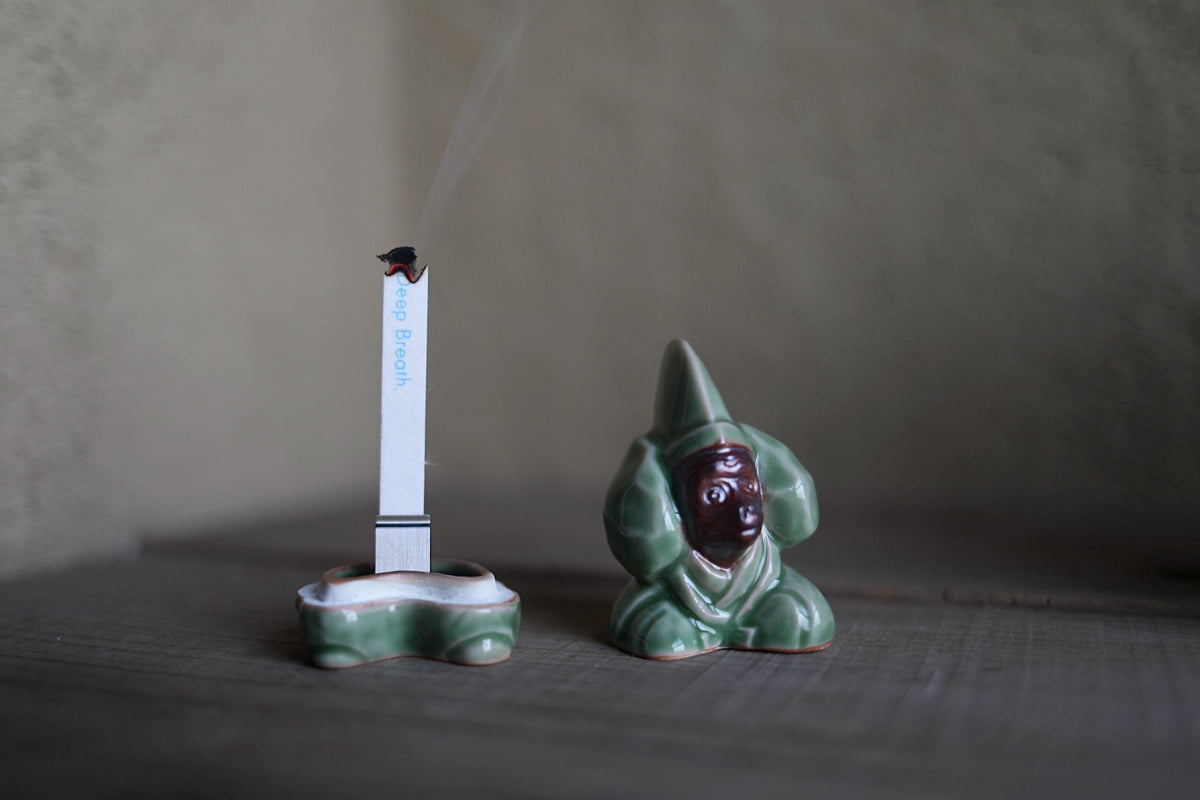 A ceramic figurine of a person sitting on a wooden surface, with a lit cigarette in a holder next to it.