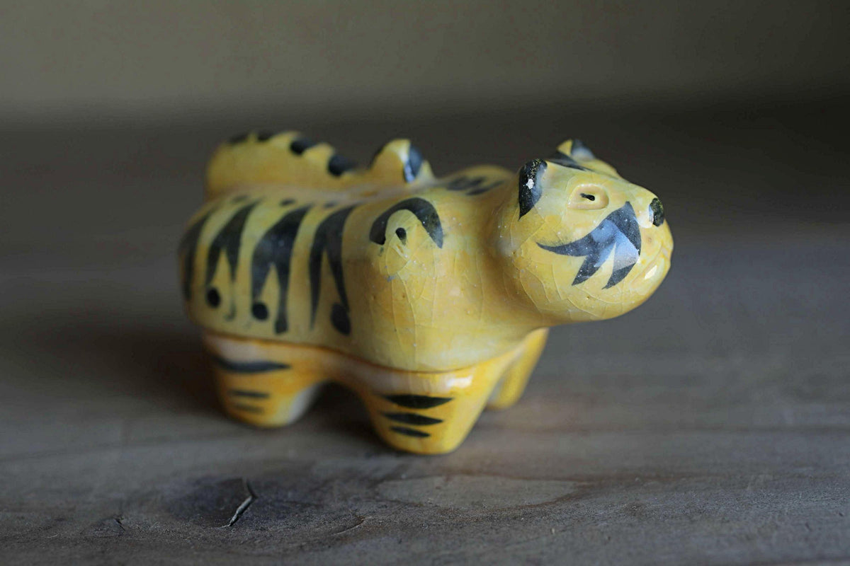 A yellow and black ceramic figurine of a tiger-like animal, possibly a cat or a small dog, sitting on a wooden surface.