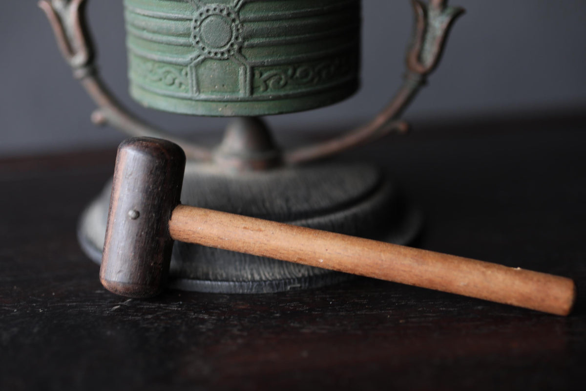 A wooden mallet rests on a dark surface, with a green metal object, possibly a bell or a trophy, visible in the background.