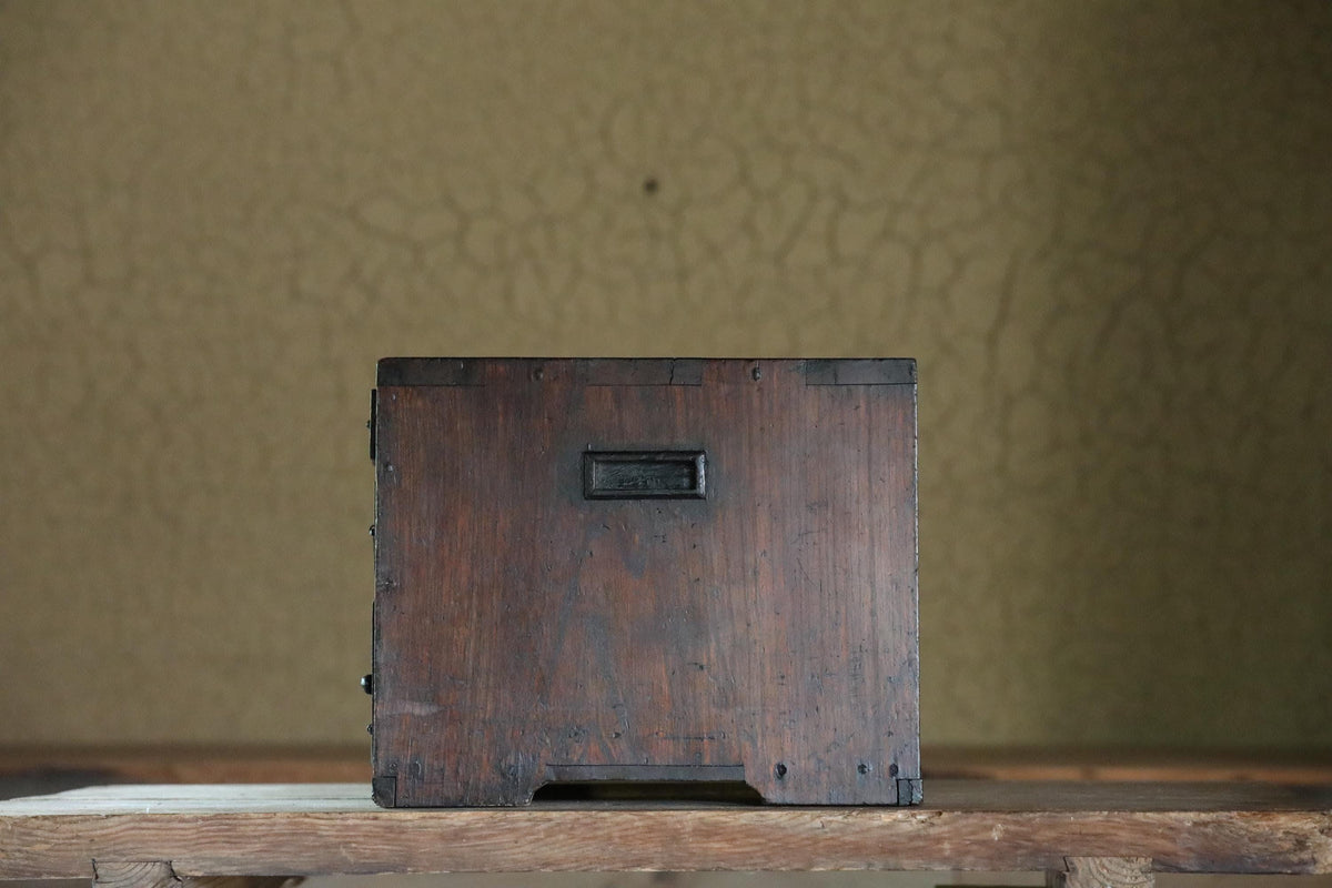 A wooden box with a metal latch on the front sits on a wooden surface against a textured green wall.