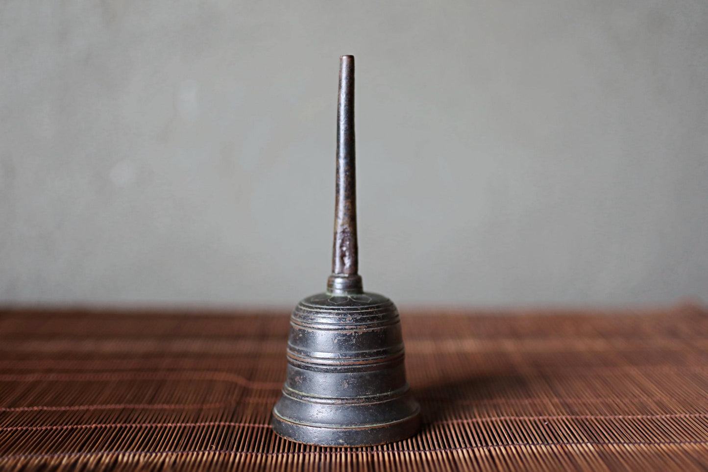 A metal bell with a long, thin stem is sitting on a brown woven mat against a gray background.