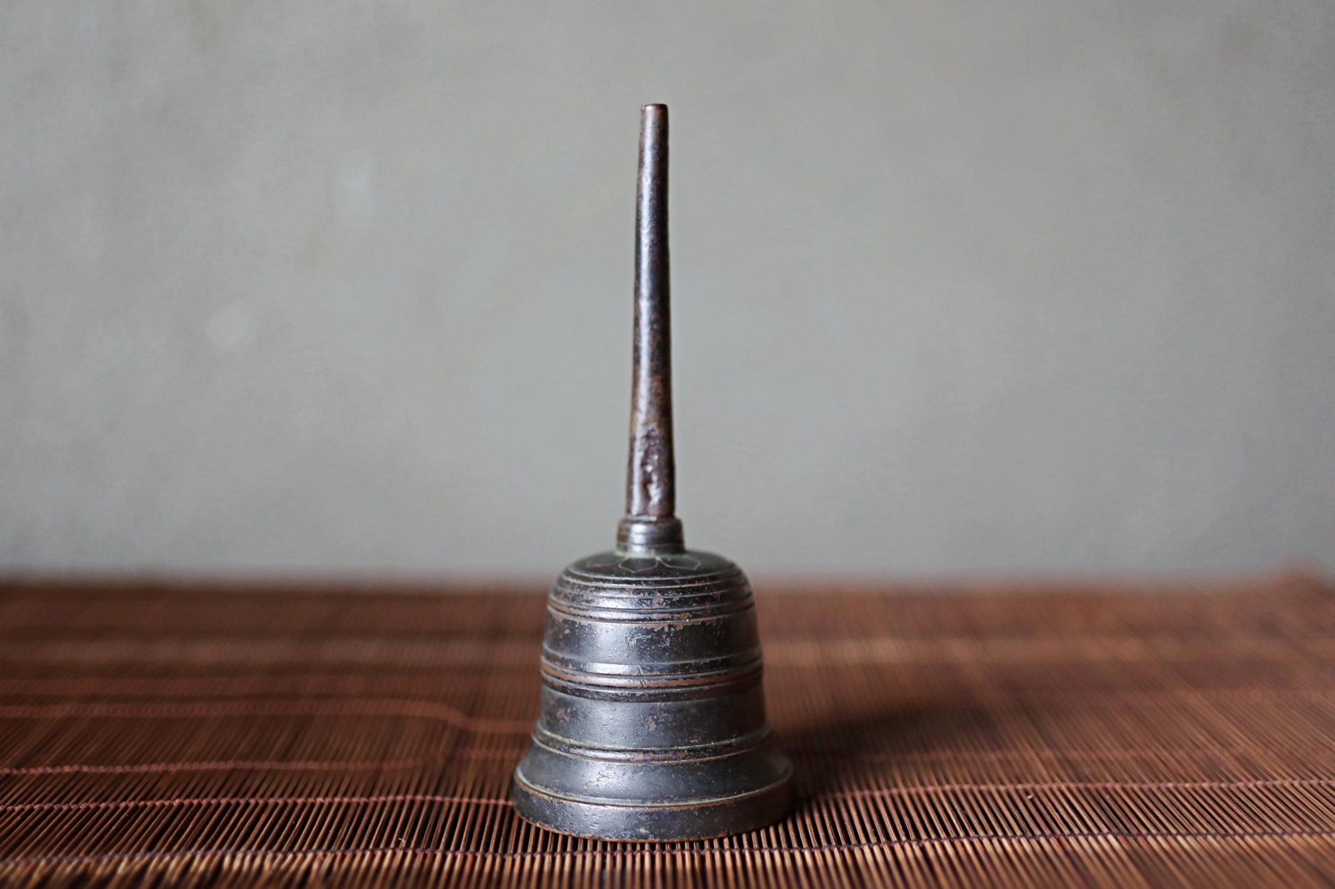 A metal bell with a long, thin stem is sitting on a brown woven mat against a gray background.