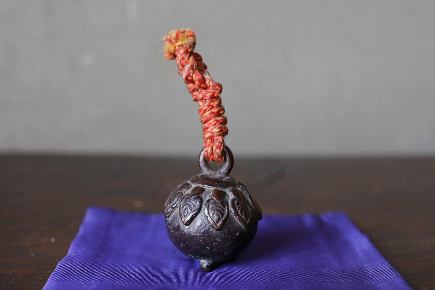 A small, ornate metal bell with a red string hanging from it, placed on a purple cloth.