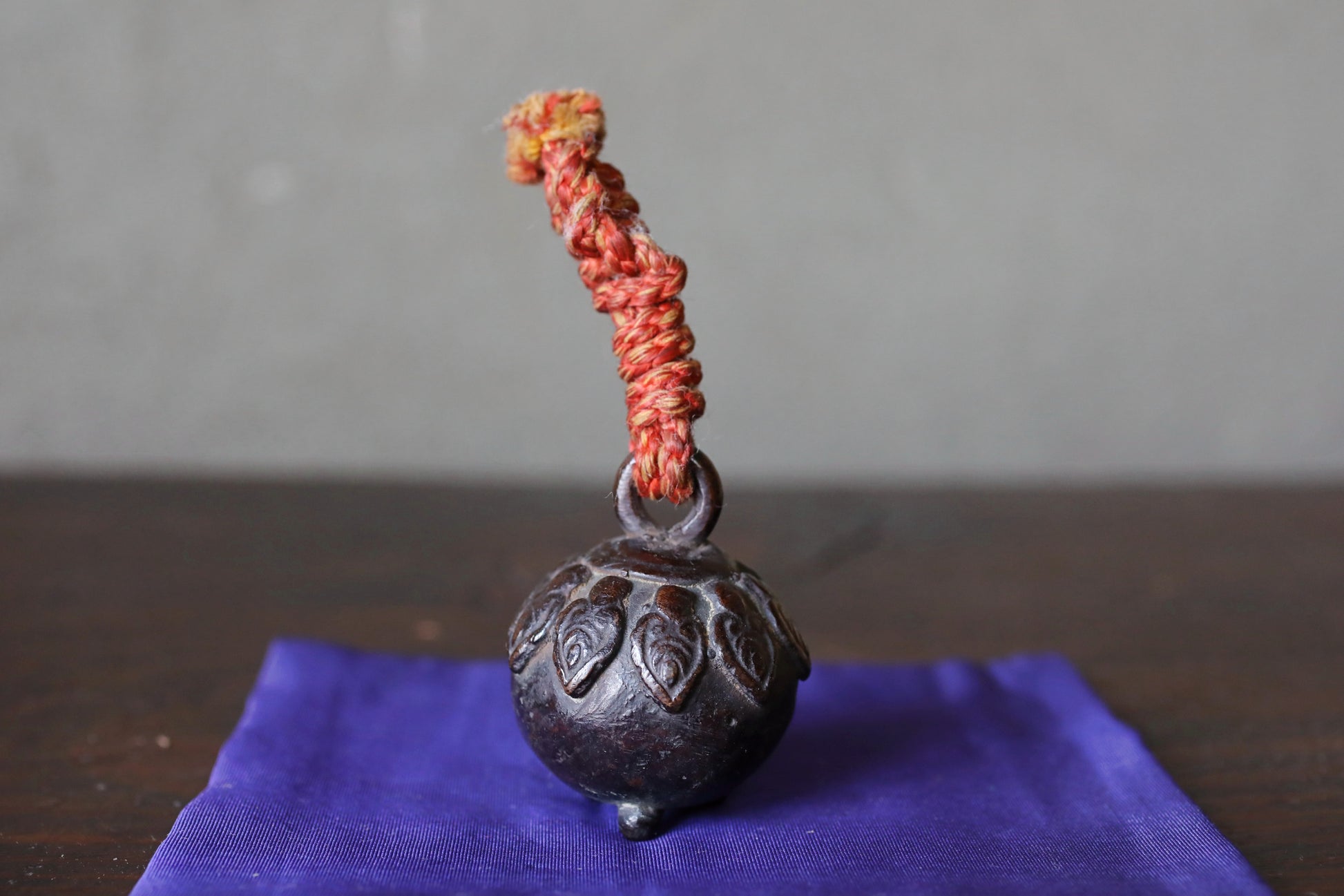A small, ornate metal bell with a red string hanging from it, placed on a purple cloth.