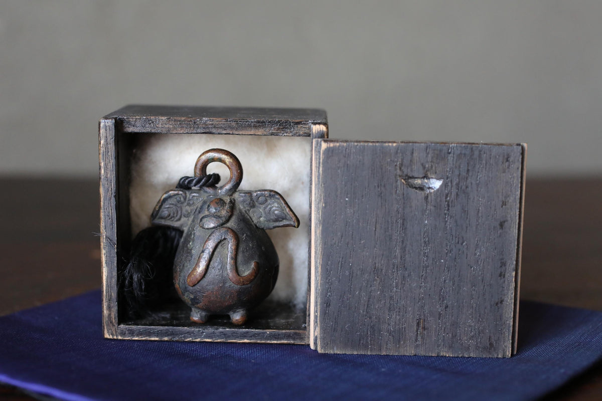 A small, ornate metal object, possibly a decorative piece, is displayed inside a wooden box on a blue cloth.
