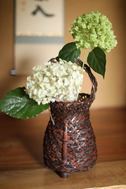 A woven basket containing white hydrangeas and a green hydrangea flower, placed on a wooden surface.