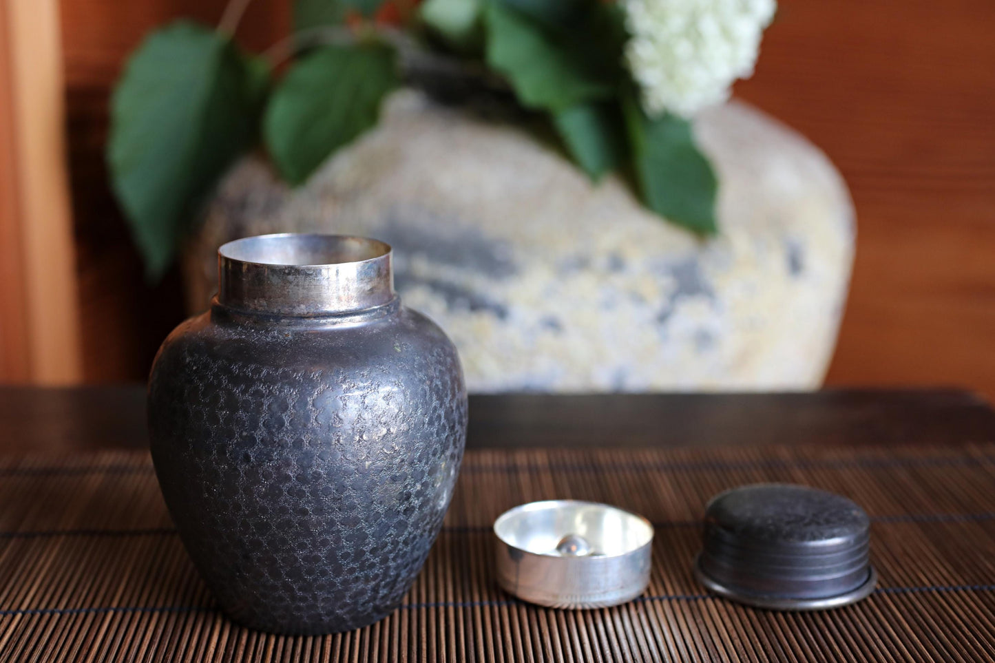 A black metal urn with a silver lid sits on a bamboo mat, with a white flower in the background.