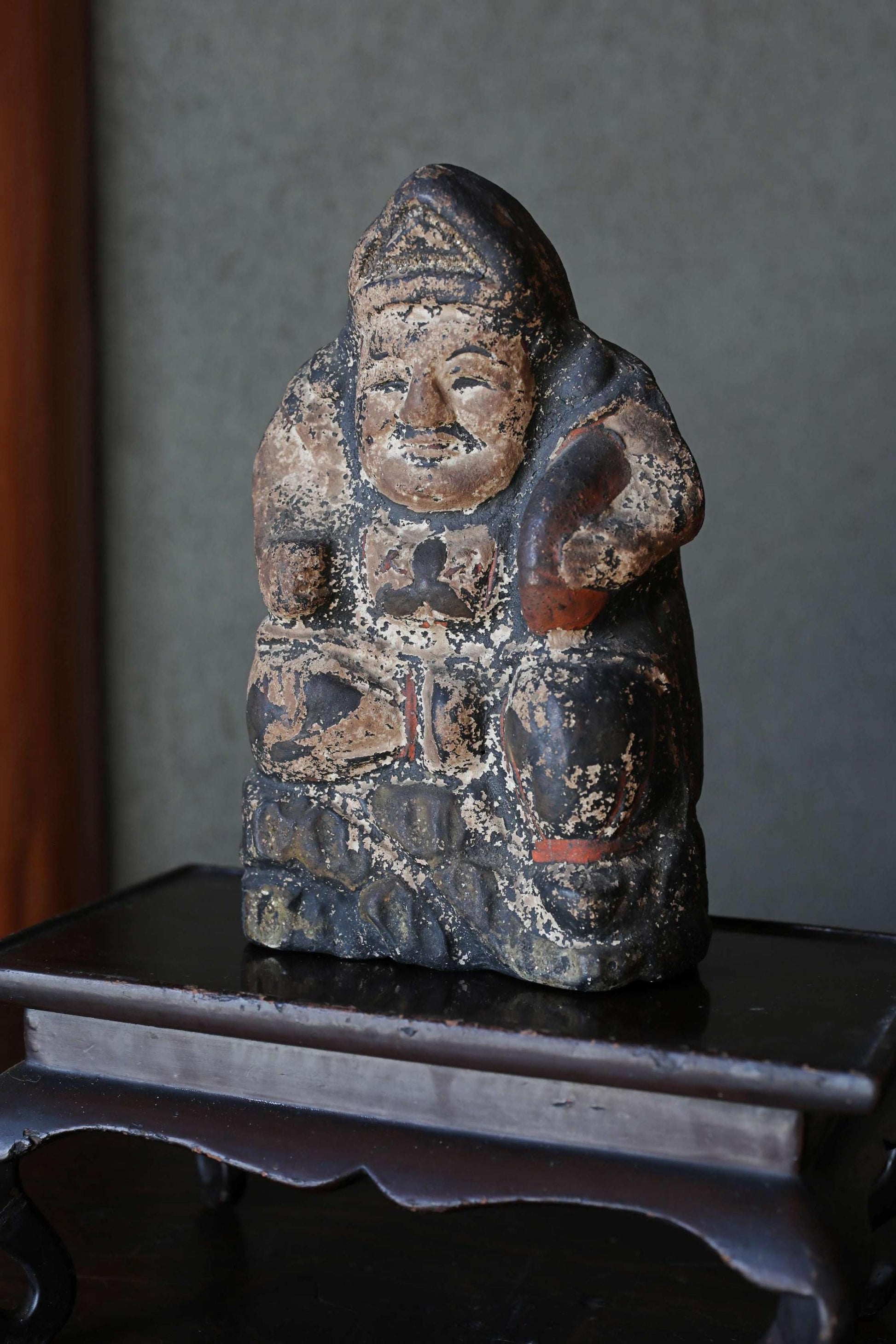 An old, weathered statue of a seated figure, possibly a deity or religious figure, displayed on a wooden stand.