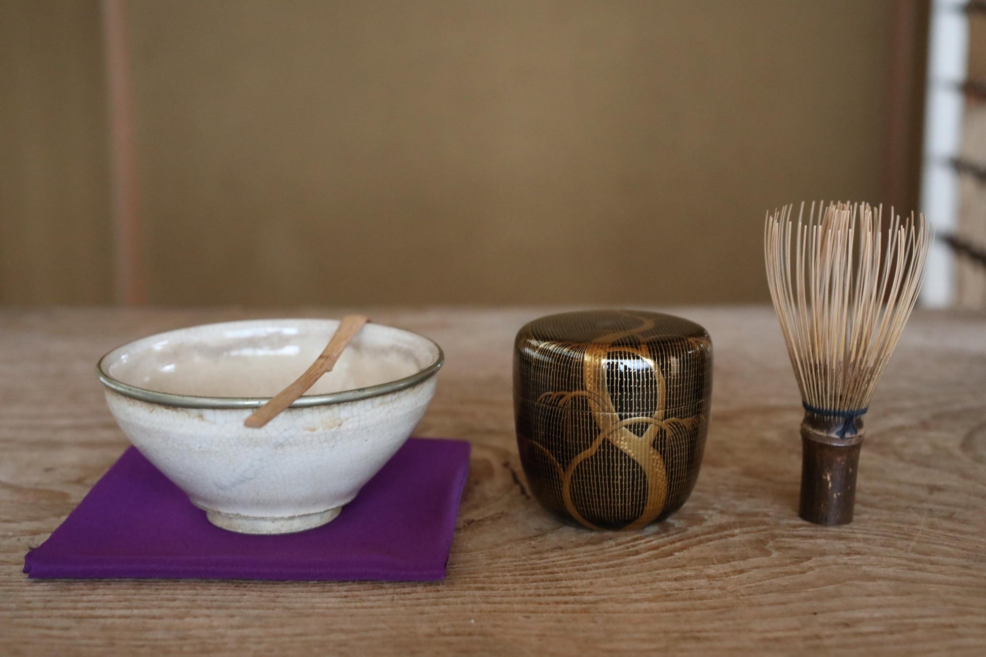 A white bowl with a wooden spoon, a black and gold ceramic container, and a wooden whisk are arranged on a purple cloth on a wooden table.
