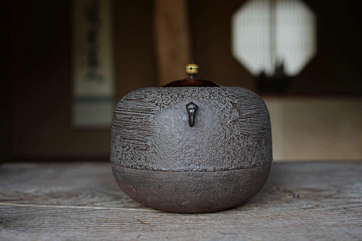 A dark brown ceramic teapot with a small handle and a small yellow ball on top, sitting on a wooden surface.