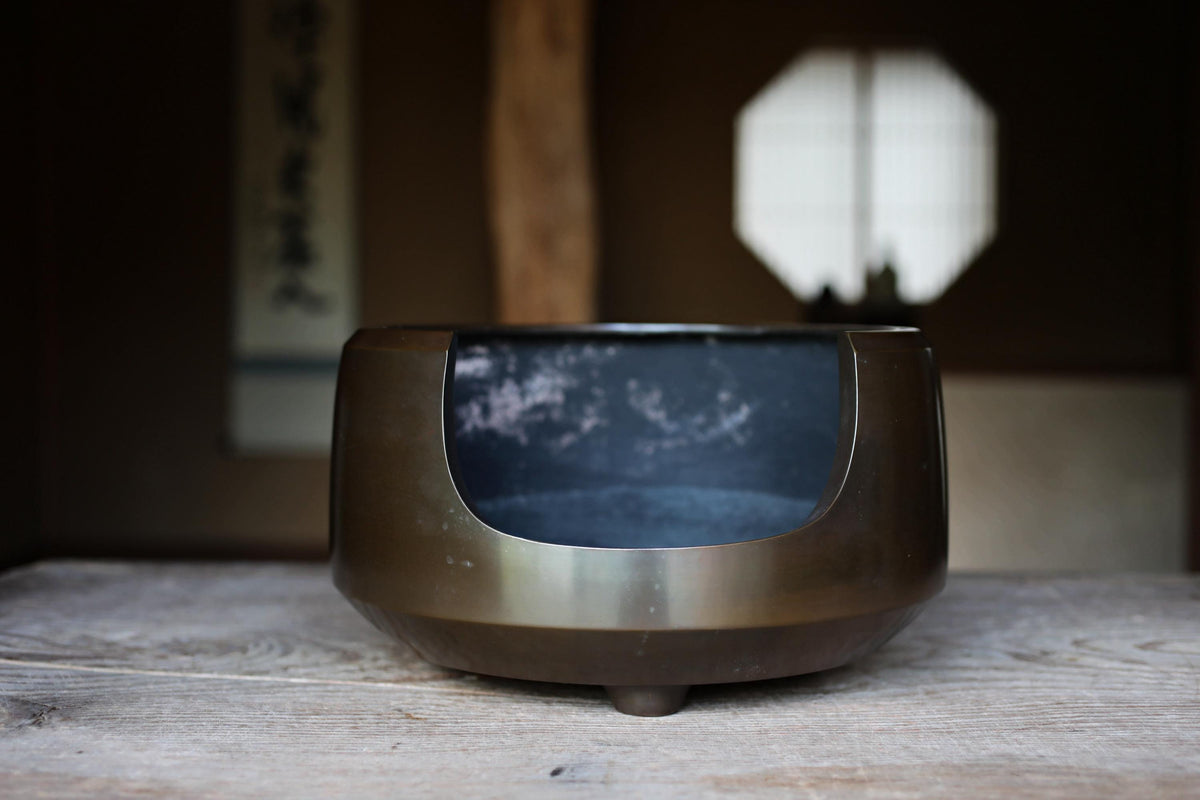 A dark metal bowl with a curved shape sits on a wooden surface, with a window and a wall in the background.