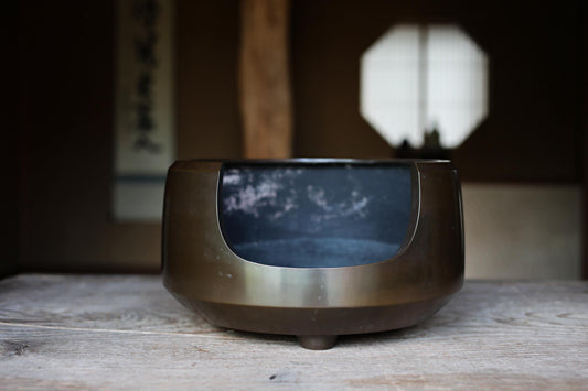 A dark metal bowl with a curved shape sits on a wooden surface, with a window and a wall in the background.