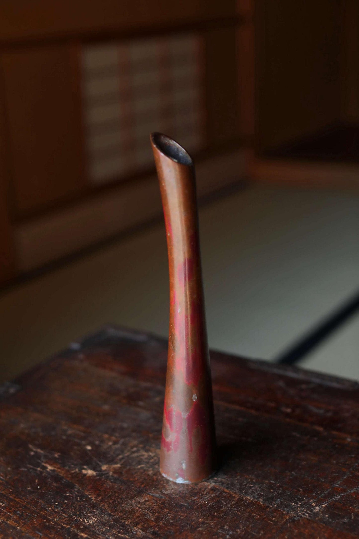 A tall, narrow vase with a reddish-brown hue sits on a wooden surface.