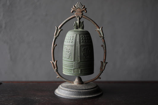 A green metal bell with a decorative metal frame is displayed on a dark surface.