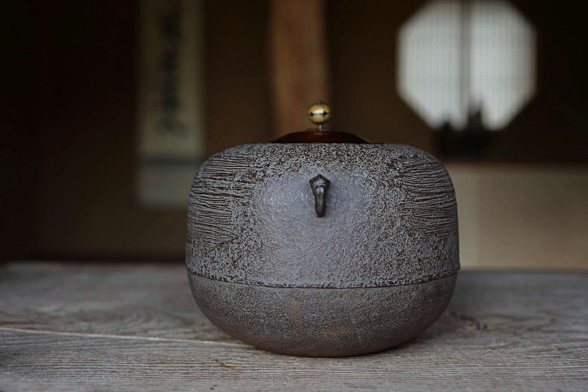 A ceramic tea kettle with a textured surface sits on a wooden surface, with a window and a wall in the background.