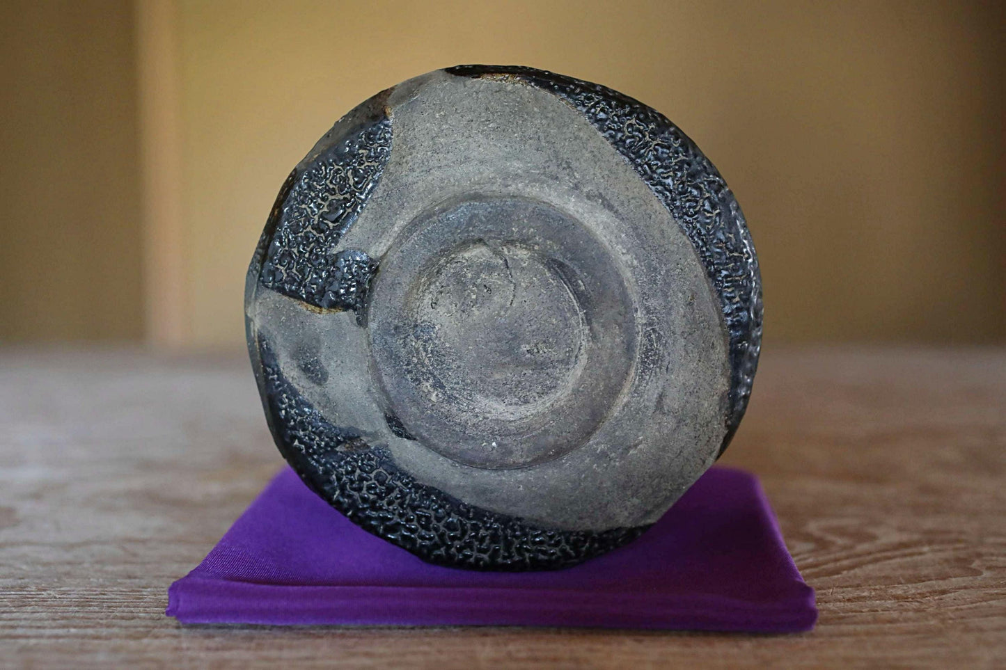 a black and gray stone or ceramic object, possibly a vase or bowl, placed on a purple cloth or cushion on a wooden surface.