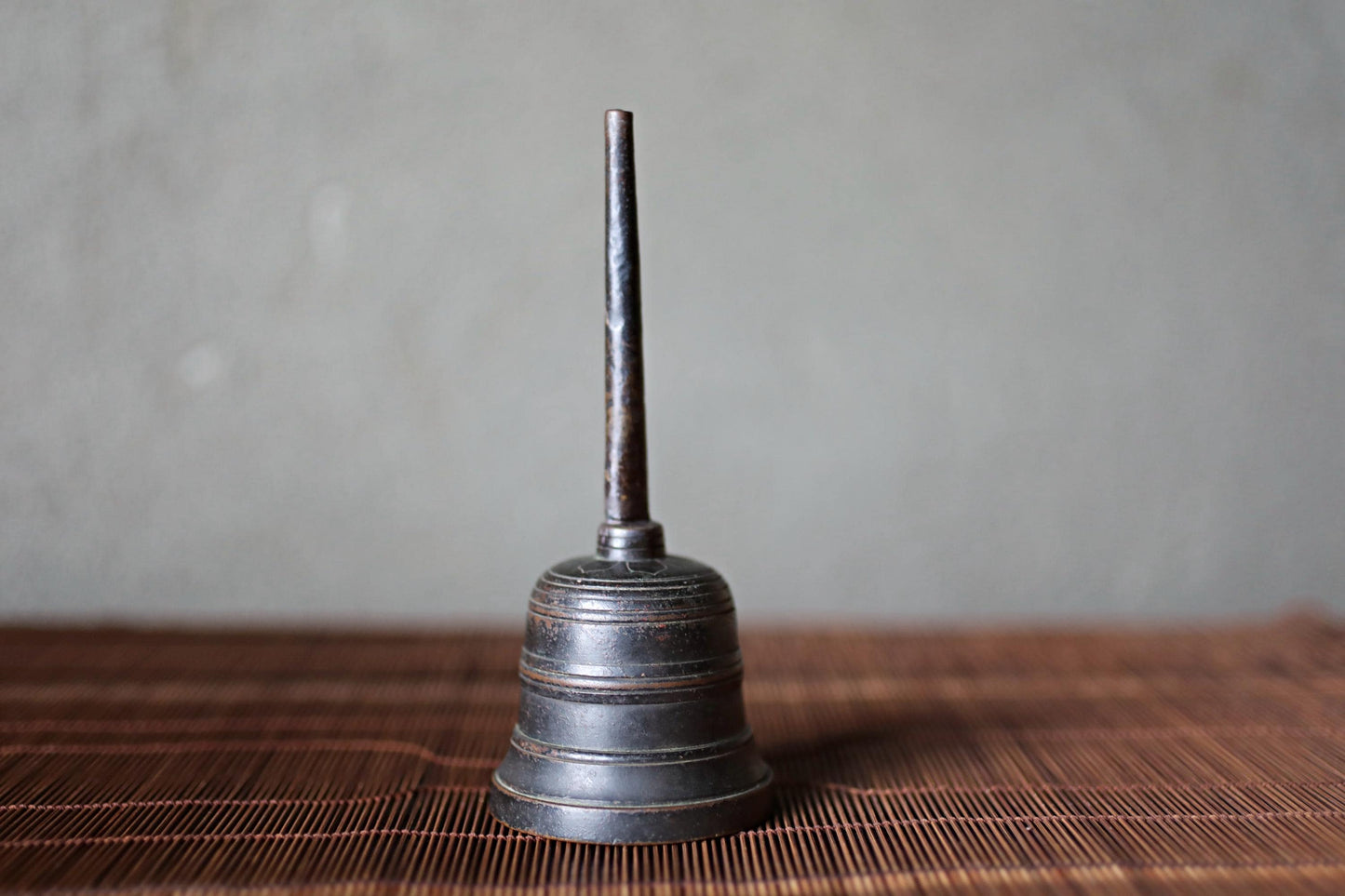 A metal bell with a pointed end is sitting on a woven mat against a plain background.